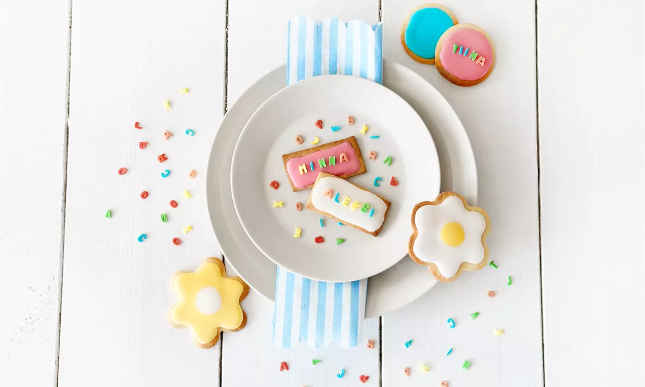 Pastellinväriset pikkuleivät nimikointikoristeilla aseteltuna valkoiselle lautaselle siniraidallisen servetin päällä, ympärillä pieniä värikkäitä strösseleitä