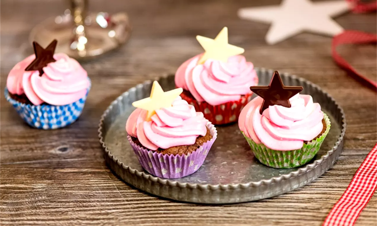 Vaaleanpunaisella kuorrutteella viimeistellyt, paperivuokiin asetetut cupcake-leivokset, joiden huipulla suklaa- ja valkotähtikoristeet, metallitarjottimella