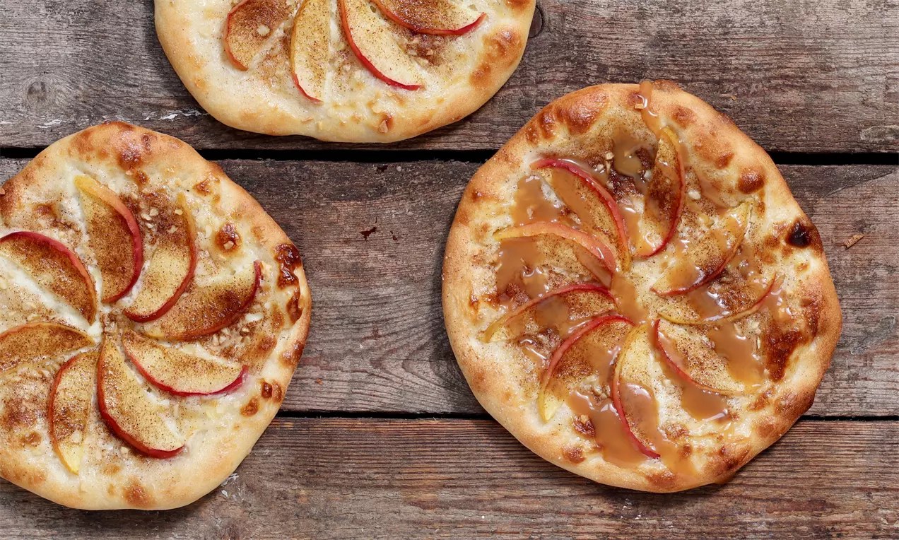Ohut pyöreä pizzapohja, päällä karamellikastiketta, kanelia ja viuhkamaisesti aseteltuja punaisia omenaviipaleita, puisen alustan päällä