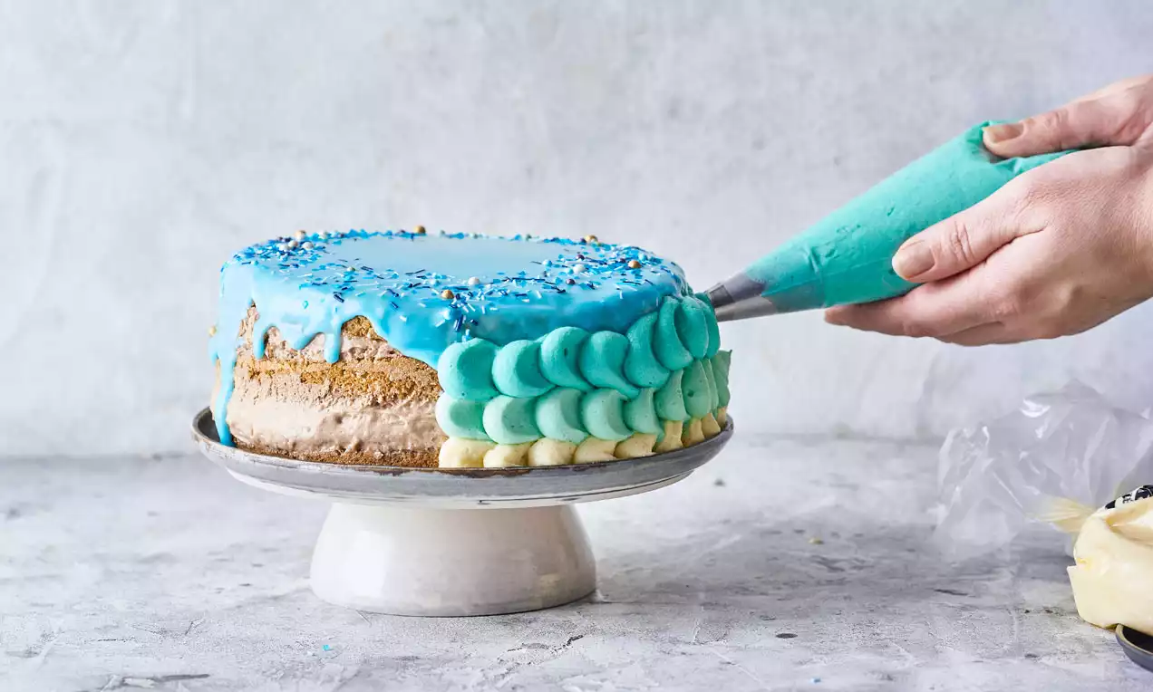 Vaaleansinistä kuorrutetta valuva pyöreä kakku matalalla jalustalla, reunaan syntyy kerroksellinen kuviopursotus pastellisessa sävyssä