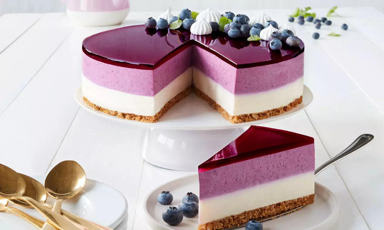 Liilalla kiilteellä kuorrutettu kerroskakku, jossa valkoinen ja violetti mousse keksipohjan päällä, koristeena mustikoita ja pieniä marenkeja