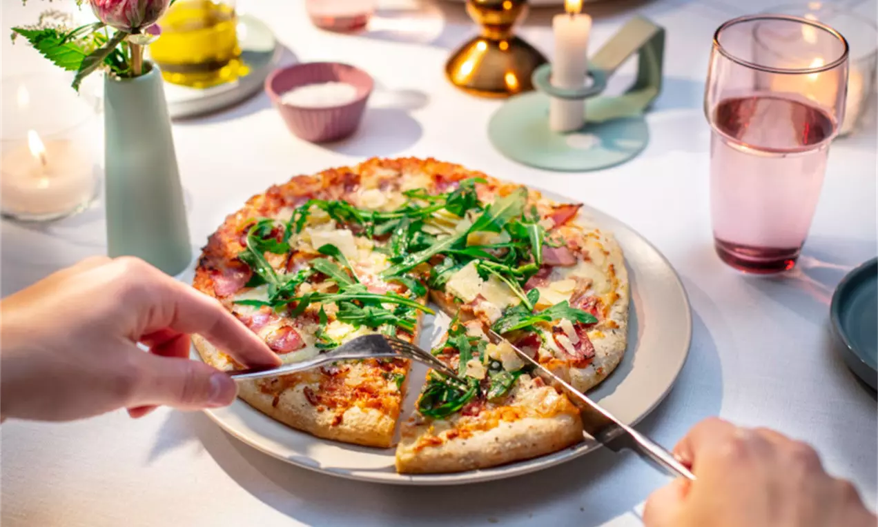 Ohut pohjainen pizza rapeilla reunoilla, täytetty vihreällä rucolalla, sulalla juustolla ja kinkulla, aseteltuna vaalealle lautaselle kynttilöiden valossa