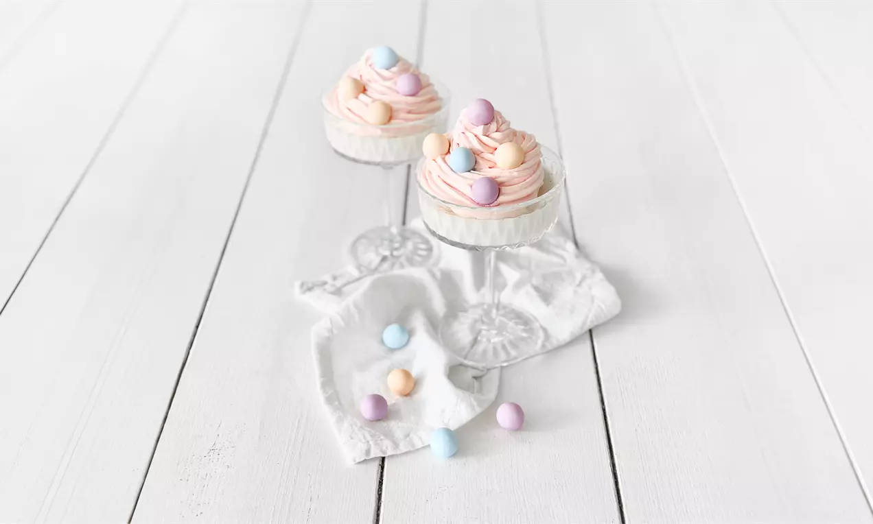 Pastellinväriset valkosuklaapannacotat kevyissä lasimaljoissa, pehmeän kermaiset pinnat koristeltuna vaaleilla sokerikuorrutetuilla suklaapalloilla