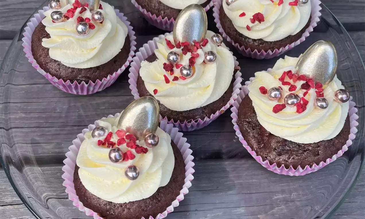 Tummanruskea cupcake vaaleaa kermavaahtoa muistuttavaa kuorrutetta, hopeista mantelia ja punaisia murusia lasilautasella
