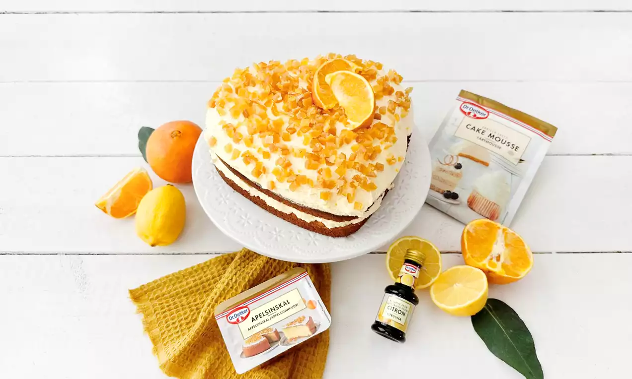 Pyöreä, kahden kerroksen ambrosiakakku valkoisella lautasella, kukkuroillaan kermavaahtoa ja koristeltu oranssinkeltaisilla hedelmänmuruilla