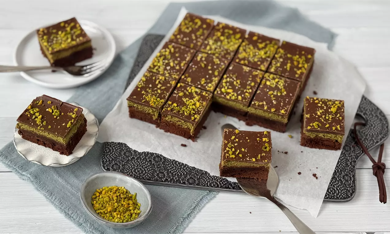 Neliönmuotoiset suklaiset brownies-leivokset, tasaisen tumman kuorrutteen ja vihreän pistaasirouheen peittämät, aseteltu vaalealle lautaselle